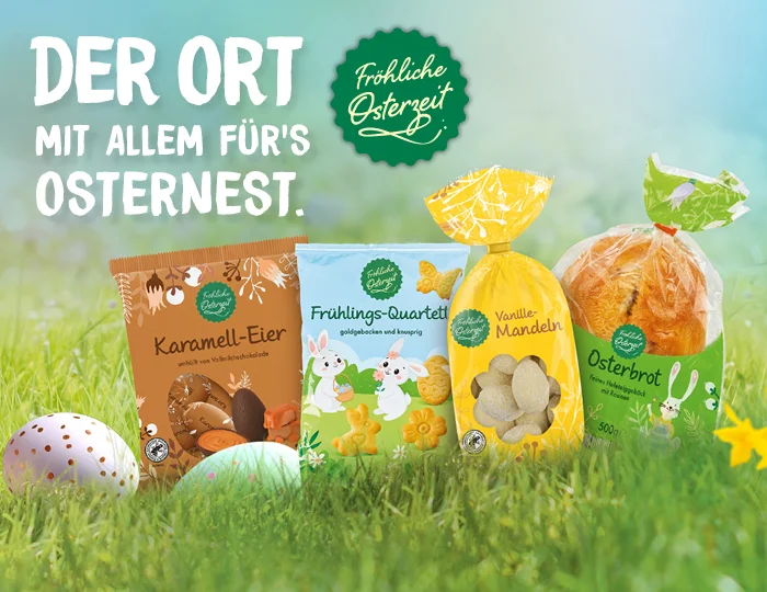 Verschiedene Osterprodukte stehen auf einer gr&uuml;nen Wiese mit Ostereiern und Fr&uuml;hlingsblumen. Zu sehen sind Karamell-Eier, Fr&uuml;hlings-Quartett Geb&auml;ck, Vanille-Mandeln und Osterbrot, jeweils in bunter Verpackung. Oben links steht in gro&szlig;en wei&szlig;en Buchstaben: &bdquo;Der Ort mit allem f&uuml;r&rsquo;s Osternest.&ldquo; Daneben ein gr&uuml;nes Siegel mit der Aufschrift &bdquo;Fr&ouml;hliche Osterzeit&ldquo;. Am unteren Rand befindet sich ein wei&szlig;er Button mit rotem Rand und der Aufschrift &bdquo;Jetzt Angebote entdecken&ldquo;, der zum Klicken einl&auml;dt.