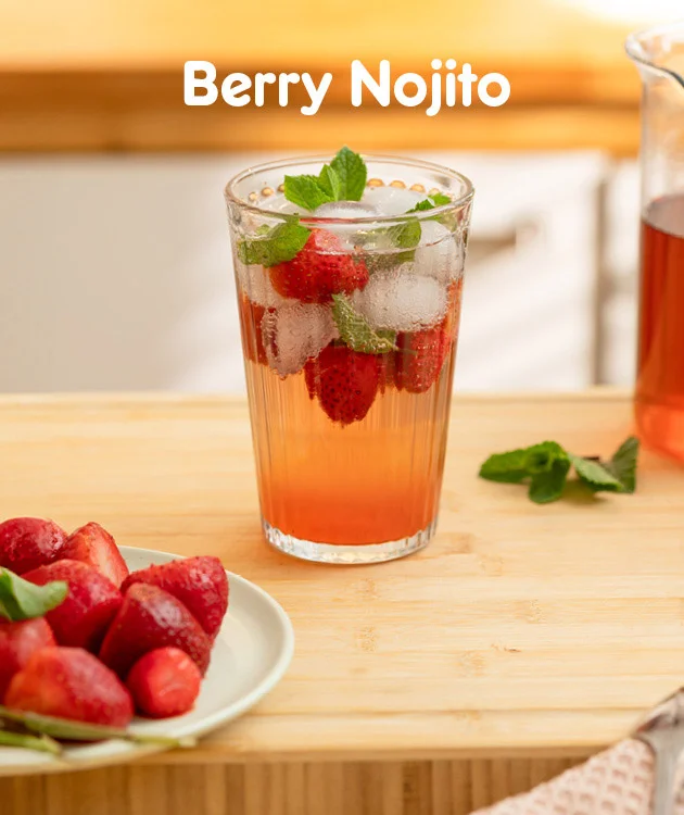 Die Grafik zeigt ein Glas mit Berry Nojito, gef&uuml;llt mit Eisw&uuml;rfeln, frischen Erdbeeren und Minzbl&auml;ttern auf einem hellen Holztisch. Im Hintergrund steht eine Karaffe mit diesem Getr&auml;nk und daneben liegen weitere Minzbl&auml;tter. Links unten ist eine Schale mit frischen Erdbeeren zu sehen. Oben steht in wei&szlig;er Schrift &bdquo;Berry Nojito&ldquo;. Unten befindet sich ein wei&szlig;-roter Button mit der Aufschrift &bdquo;Hier Rezept entdecken&ldquo;, der zum Klicken einl&auml;dt.