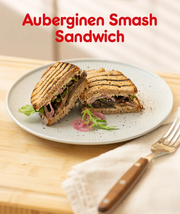 Auf einem hellen Teller liegen zwei H&auml;lften eines Auberginen Smash Sandwichs mit Grillstreifen, gef&uuml;llt mit Auberginen, Salat und roten Zwiebeln. Im Hintergrund liegt Besteck. Oben steht in gro&szlig;er roter Schrift &bdquo;Auberginen Smash Sandwich&ldquo;. Unten befindet sich ein wei&szlig;er Button mit rotem Rand und roter Aufschrift &bdquo;Hier Rezept entdecken&ldquo;, der zum Klicken einl&auml;dt.
