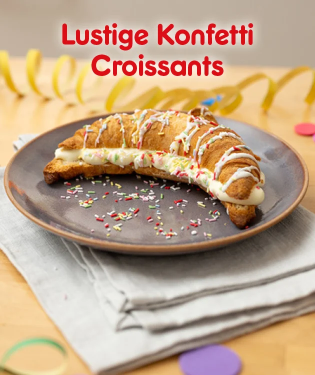 Ein Croissant liegt auf einem braunen Teller und ist mit wei&szlig;er Glasur und bunten Zuckerstreuseln dekoriert. Im Hintergrund liegen gelbe Luftschlangen und bunte Konfetti auf einem Holztisch. Oben steht in gro&szlig;en roten Buchstaben: &bdquo;Lustige Konfetti Croissants&ldquo;. Am unteren Rand befindet sich ein wei&szlig;er Button mit rotem Rand und der Aufschrift &bdquo;Hier Rezept entdecken&ldquo;, der zum Klicken einl&auml;dt.
