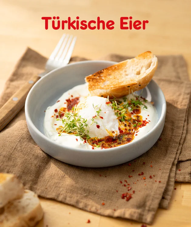 Die Grafik zeigt einen hellen Holzhintergrund, auf dem eine Schale mit &bdquo;T&uuml;rkische Eier&ldquo; steht. In der grauen Sch&uuml;ssel sieht man pochierte Eier auf cremigem Joghurt, betr&auml;ufelt mit w&uuml;rzigem Chili-&Ouml;l und bestreut mit frischen Kr&auml;utern. Daneben liegt eine ger&ouml;stete Brotscheibe am Rand der Schale, im Hintergrund befinden sich ein Holzbrett mit Gabel sowie ein Leinentuch.