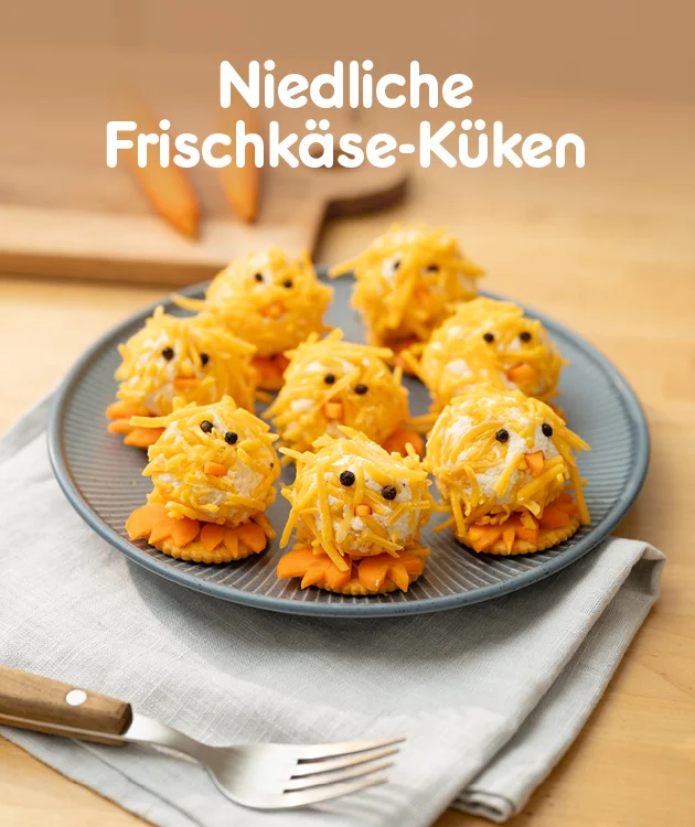 Grafik mit der &Uuml;berschrift &bdquo;Niedliche Frischk&auml;se-K&uuml;ken&ldquo;. In der Mitte steht ein Teller mit mehreren gelben Frischk&auml;se-K&uuml;ken aus geriebenem K&auml;se, mit Augen und Schnabel, jeweils auf orangefarbenen Crackern, auf einem Tuch auf einem Holztisch. Unten ein Button &bdquo;Hier Rezept entdecken&ldquo;, der zum Klicken einl&auml;dt.