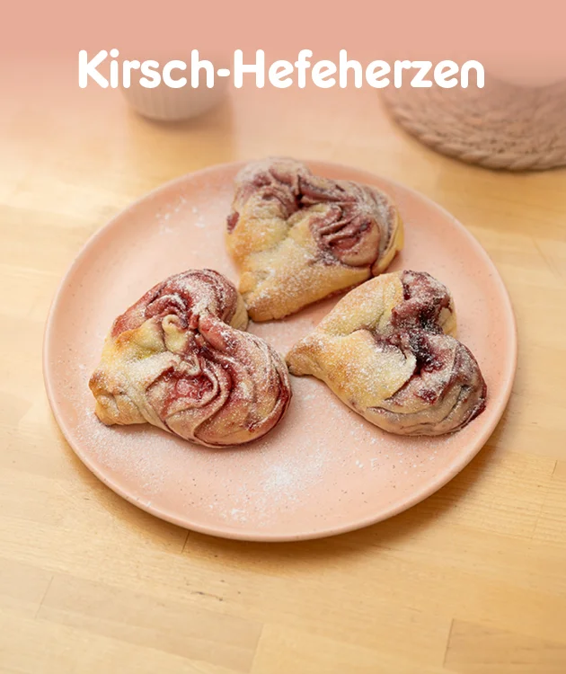 Diese Grafik zeigt einen hellen Holztisch mit einem rosa Teller, darauf liegen drei Kirsch-Hefeherzen mit sichtbarer Kirschf&uuml;llung. Oben steht als Headline &bdquo;Kirsch-Hefeherzen&ldquo;. Ganz unten ist ein Button &bdquo;Hier Rezept entdecken&ldquo;, der zum Klicken einl&auml;dt.