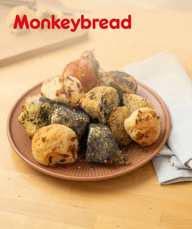 Gezeigt wird ein Rezeptmotiv f&uuml;r &bdquo;Monkeybread&ldquo;. Oben links steht der Titel in roter Schrift. In der Mitte ist ein Teller mit mehreren goldbraunen und teils dunkel bestreuten Geb&auml;ckst&uuml;cken auf einem Holztisch zu sehen. Rechts daneben liegt eine gefaltete Stoffserviette. Unten befindet sich ein Button mit der Aufschrift &bdquo;Hier Rezept entdecken&ldquo;, der zum Klicken einl&auml;dt.