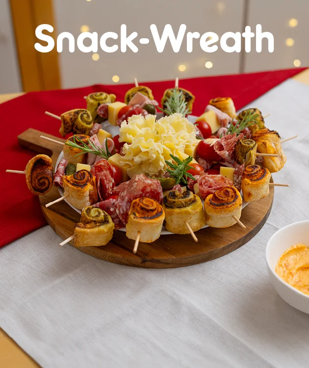 Bild mit festlich gedecktem Tisch, im Hintergrund Lichter. Oben mittig weiße Schrift: „Snack-Wreath“. Im Vordergrund ein rundes Holzbrett mit einem Snack-Kranz aus herzhaften Spießen, belegt mit Blätterteigschnecken, Käse, Salami, Schinken, Tomaten und Kräutern, in der Mitte dekorativ geriebener Käse und Rosmarinzweige. Unten mittig ein weiß-roter Button mit der Aufschrift: „Hier Rezept entdecken“, der zum Klicken einlädt.