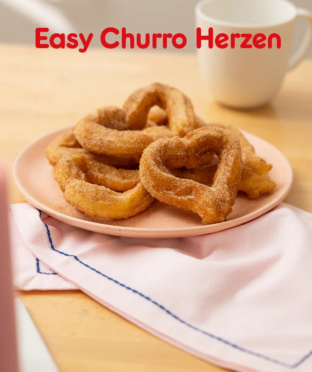 Grafik mit einem hellen, freundlichen Hintergrund. Oben steht in gro&szlig;en roten Buchstaben: &bdquo;Easy Churro Herzen&ldquo;. Im Mittelpunkt befindet sich ein rosa Teller mit mehreren herzf&ouml;rmigen Churros, die mit Zimt und Zucker bestreut sind. Im Hintergrund ist eine wei&szlig;e Tasse zu sehen. Der Teller steht auf einer hellrosa Serviette mit blauer Ziernaht. Unten mittig befindet sich ein wei&szlig;er Button mit rotem Rand und roter Schrift: &bdquo;Hier Rezept entdecken&ldquo;, der zum Klicken einl&auml;dt.