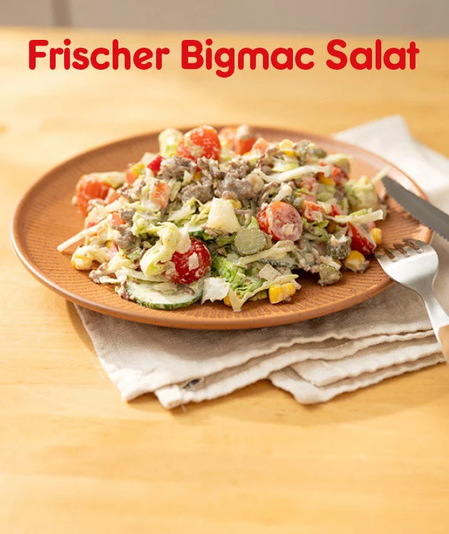 Grafik mit einem braunen Teller, darauf ein frischer Bigmac Salat mit Zutaten wie Salat, Tomaten, Gurken, Mais, Hackfleisch und K&auml;se, angerichtet auf einem Holztisch mit beiger Serviette, Messer und Gabel rechts daneben. Oben in gro&szlig;er roter Schrift: &bdquo;Frischer Bigmac Salat&ldquo;. Unten mittig ein wei&szlig;er Button mit rotem Rand und roter Schrift: &bdquo;Hier Rezept entdecken&ldquo;, der zum Klicken einl&auml;dt.