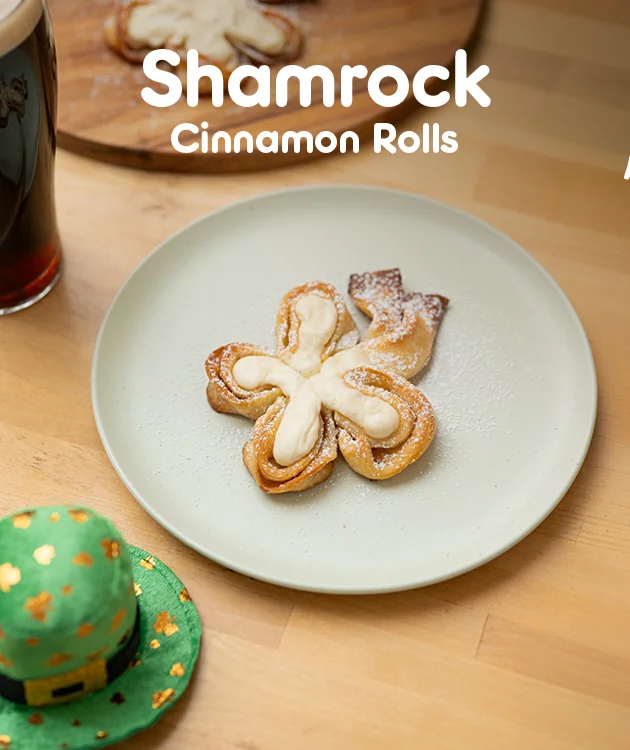 Die Grafik zeigt einen Teller mit Shamrock Cinnamon Rolls auf einem Holztisch neben einem Getr&auml;nk, mit der Aufschrift &bdquo;Shamrock Cinnamon Rolls&ldquo; und dem Button &bdquo;Hier Rezept entdecken&ldquo;.