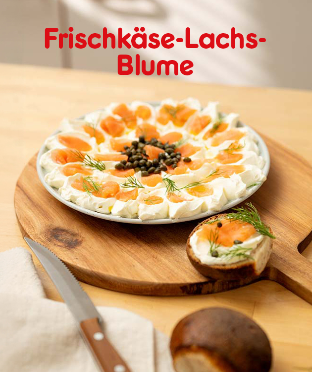 Motiv mit der &Uuml;berschrift &bdquo;Frischk&auml;se-Lachs-Blume&ldquo;. Auf einem Holzbrett steht ein runder Teller mit einer blumenf&ouml;rmig angerichteten Frischk&auml;secreme, belegt mit Lachs, Kr&auml;utern und Kapern in der Mitte. Rechts unten ein St&uuml;ck Brot mit Frischk&auml;se und Lachs. Unten mittig Button &bdquo;Hier Rezept entdecken&ldquo;; der Button l&auml;dt zum Klicken ein.