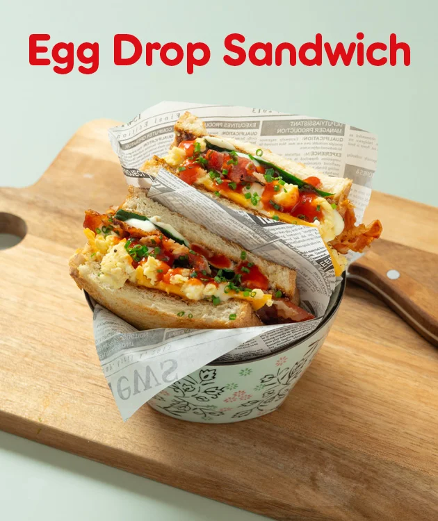 Diese Grafik zeigt den Schriftzug &bdquo;Egg Drop Sandwich&ldquo;, zwei belegte Sandwiches in Papier eingeschlagen in einem Becher auf einem Holzbrett sowie ein Messer daneben. Unten l&auml;dt der Button &bdquo;Hier Rezept entdecken&ldquo; zum Klicken ein.